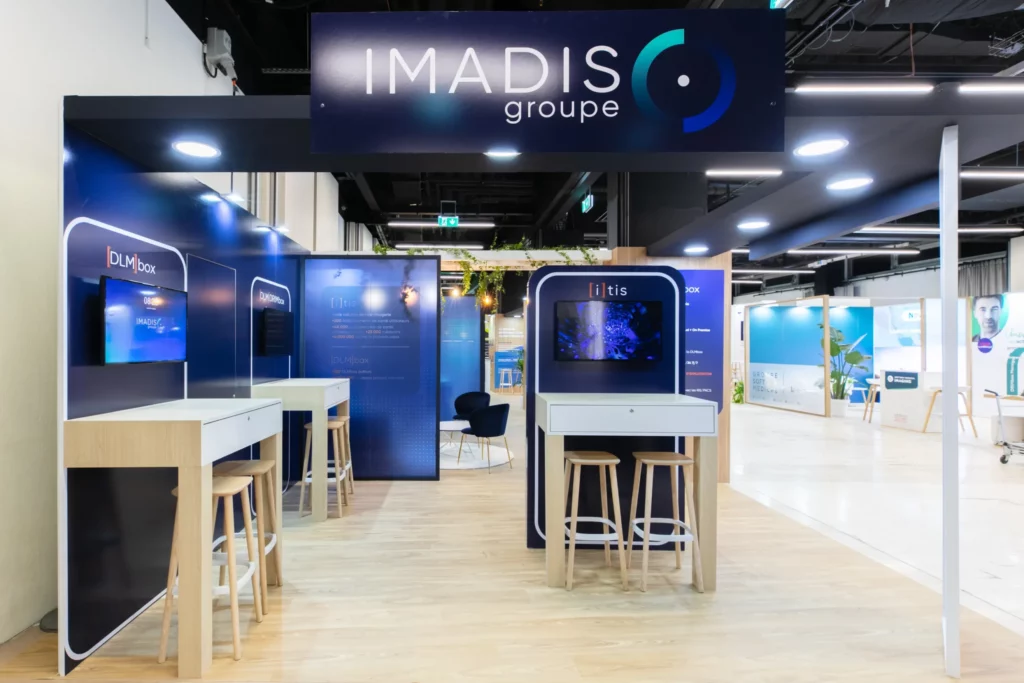 Photo de l'espace d&eacute;mo du stand IMADIS Groupe avec des consoles et &eacute;crans TV, des tabourets