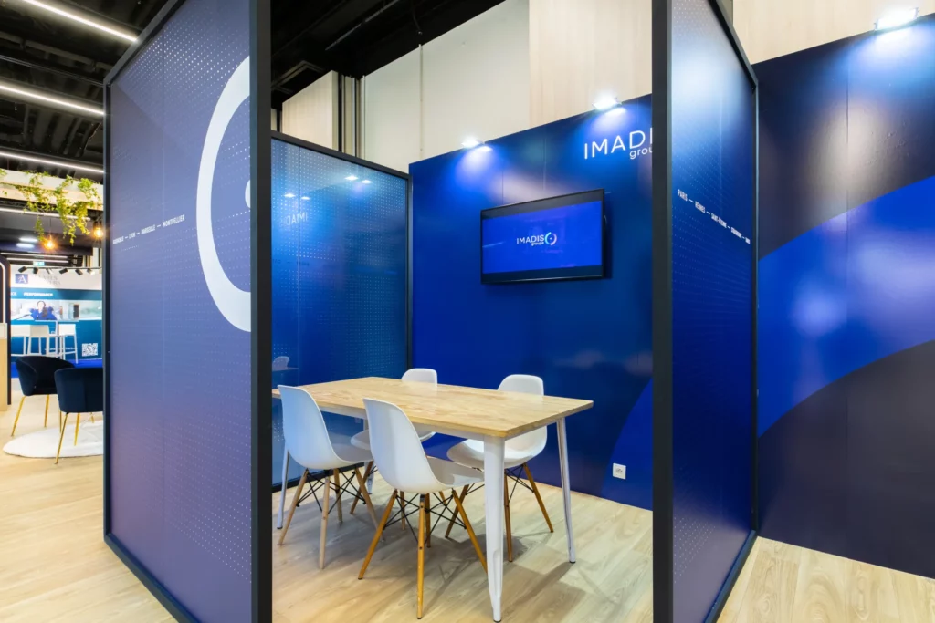 Photo d'un espace vitr&eacute; du stand IMADIS Groupe avec une table en bois, quatre chaises blanches et un &eacute;cran TV