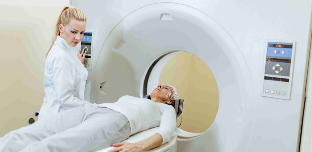 Une professionnelle de sant&eacute; accompagne une patiente allong&eacute;e sur la table d&rsquo;un scanner, illustrant la r&eacute;alisation d&rsquo;un examen d&rsquo;imagerie m&eacute;dicale dans un environnement clinique moderne.