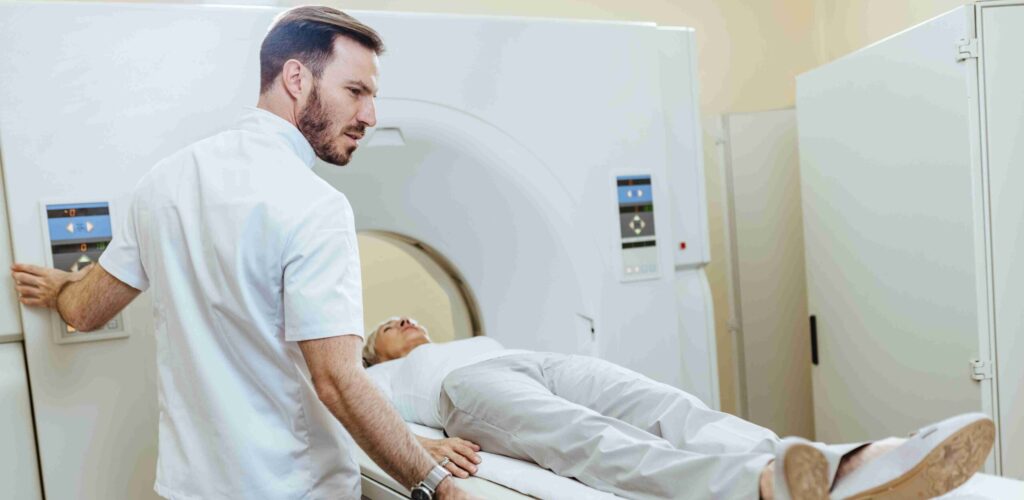 Un professionnel de sant&eacute; accompagne une patiente allong&eacute;e sur la table d&rsquo;un IRM, illustrant la r&eacute;alisation d&rsquo;un examen d&rsquo;imagerie m&eacute;dicale dans un environnement clinique moderne.